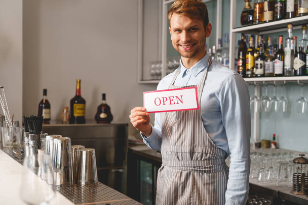 Cheerful bartender in apron holding open sign - Foto, afbeelding