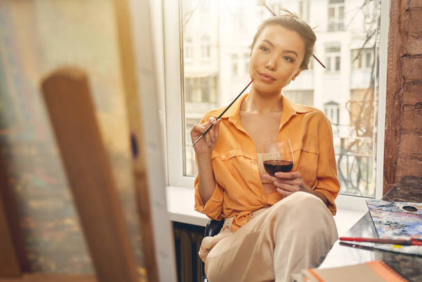 Beautiful female artist holding paintbrush and glass of wine - Foto, imagen