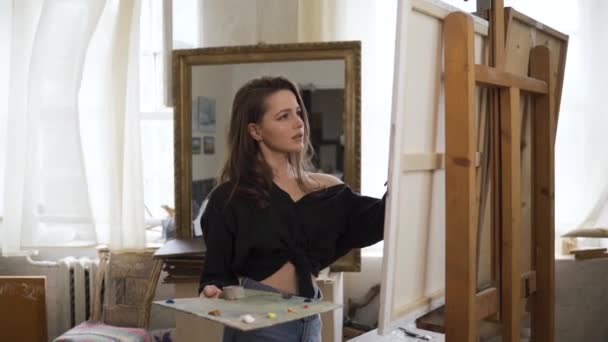A young beautiful girl artist paints a picture on an easel in the artists studio, looks into the camera and smiles. - Materiał filmowy, wideo