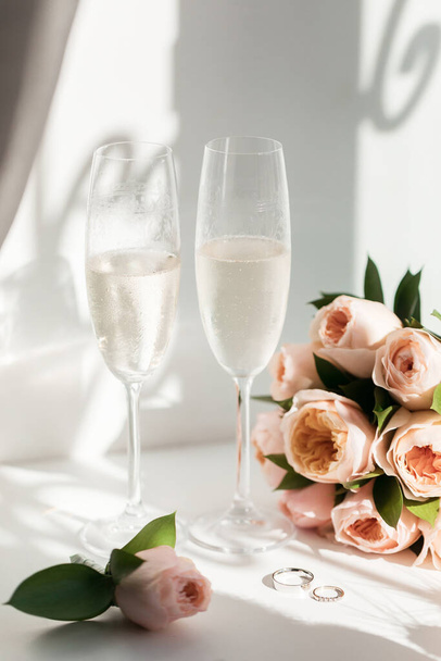 Anéis de casamento estão ao lado de dois copos de champanhe e um buquê de rosas ao sol
 - Foto, Imagem