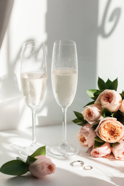 Svatební prsteny leží vedle dvou sklenic šampaňského a kytice růží na slunci - Fotografie, Obrázek