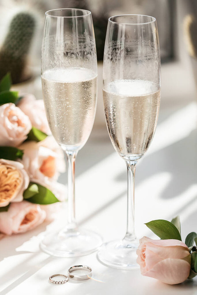 Trouwringen liggen naast twee glazen champagne en een boeket rozen in de zon - Foto, afbeelding