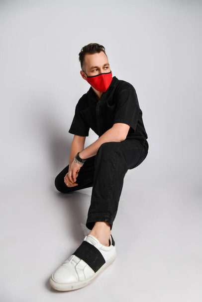 Young man in black clothing and metal accessories sits squatting in red medical mask for protection from infection - Foto, Imagem
