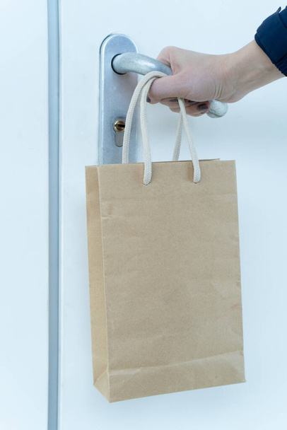 A human hand is trying to open a locked door and a paper bag of food is hanging on his wrist. Contactless food delivery. Online order of products in the store at home during the quarantine. Donate. - Фото, изображение