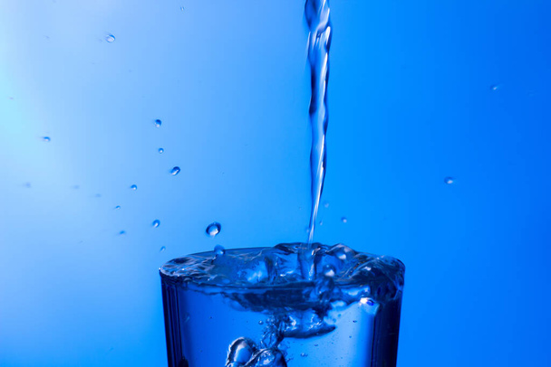 Helder drinkwater valt in een helder glas, gezond, gezond, zoet water zonder geur of smaak. Een plons waterdruppels spatten op het oppervlak. Waterstraal in het glas - Foto, afbeelding