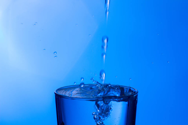 Jasne, woda pitna wpada do przezroczystej szklanki, zdrowej, zdrowej, słodkiej wody bez zapachu i smaku. Kropla wody rozpryskuje się na powierzchnię. Strumień wody wpadający do szklanki - Zdjęcie, obraz