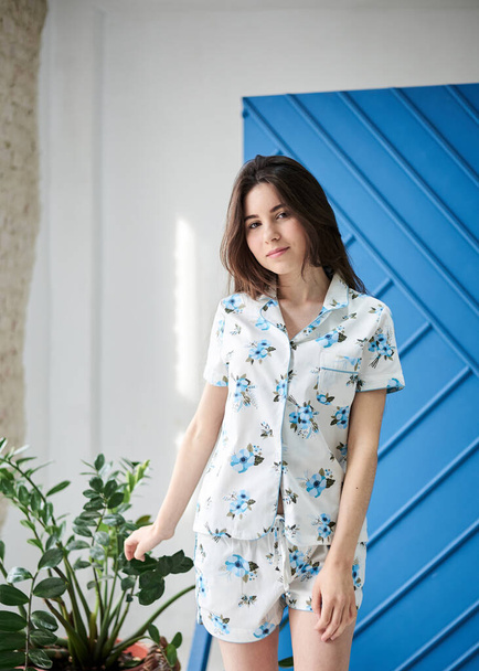 Young pretty slim brunette woman, wearing white pajamas with light blue flowers pattern, posing for picture in front of blue wall and green plant in flowerpot. Sleepwear design. Cute girl in the morning. - Fotoğraf, Görsel