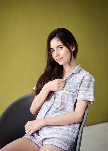 Young pretty slim brunette woman, wearing grey and white pajamas with christmas pattern, sitting in grey chair in front of light green lemon wall. Sleepwear design. Cute girl in the morning. Christmas - Φωτογραφία, εικόνα