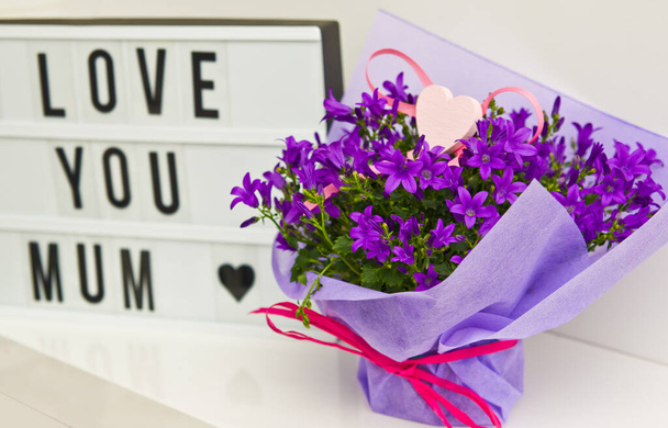 Love you mum text with campanula flowers gift wrapped in paper and ribbon on a white shelf. - 写真・画像