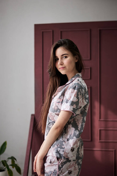 Young pretty slim brunette woman, wearing colorful light pink and grey pajamas with flowers pattern, posing for picture in front of claret marsal wall. Sleepwear design. Cute girl in the morning. - Photo, Image