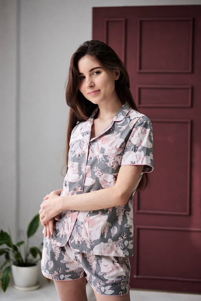 Young pretty slim brunette woman, wearing colorful light pink and grey pajamas with flowers pattern, posing for picture in front of claret marsal wall. Sleepwear design. Cute girl in the morning. - 写真・画像