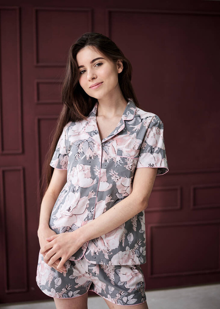 Young pretty slim brunette woman, wearing colorful light pink and grey pajamas with flowers pattern, posing for picture in front of claret marsal wall. Sleepwear design. Cute girl in the morning. - Foto, Imagem