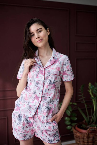 Young pretty slim brunette woman, wearing colorful light pink and grey pajamas with flowers pattern, posing for picture in front of claret marsal wall. Sleepwear design. Cute girl in the morning. - Foto, imagen