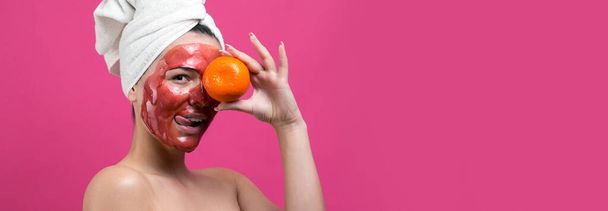 Retrato de belleza de mujer en toalla blanca en la cabeza con máscara roja nutritiva en la cara. Cuidado de la piel limpieza ecológica cosmética spa relajante concepto. Una chica está de pie con la espalda sosteniendo una mandarina naranja
. - Foto, imagen