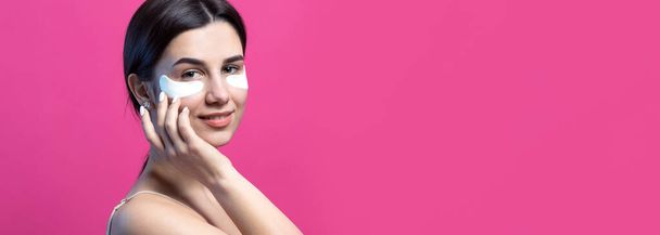 Close up portrait of pretty attractive girl with naked shoulders using patches under eyes. Standing over pink background. - Фото, изображение