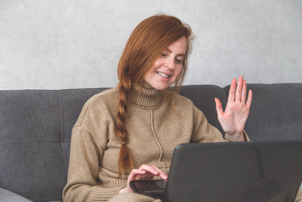 Schöne junge Frau in lässiger Kleidung benutzt einen Laptop, winkt und lächelt beim Videochat. - Foto, Bild
