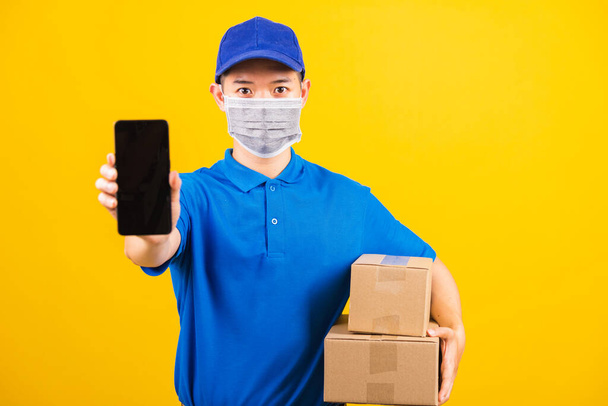 Asian young delivery worker man t-shirt and cap uniform wearing face mask protective show front black screen of smart mobile phone under coronavirus or COVID-19, studio shot isolated yellow background - Photo, Image