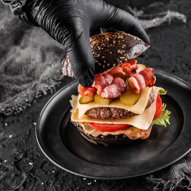 Chefs hand in black glove cooking the black burger with beaf steak, lettuce, tomatoes, slice of cheese, ham, pastrami and sauce on slate black background, close up - Photo, Image