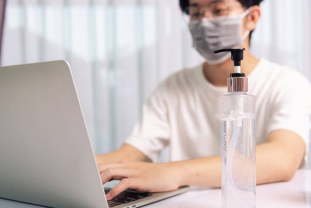 Asian business young handsome man glasses, wearing protective mask with disinfectant gel beside he work from home office remote online with laptop computer quarantines disease coronavirus or COVID-19 - Фото, изображение