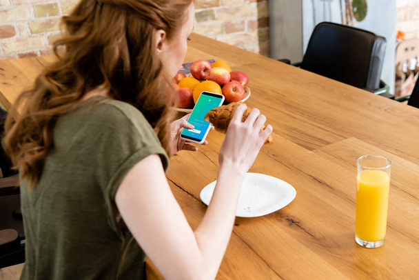 KYIV, UKRAINE - 8 mei 2020: Zijaanzicht van vrouw met smartphone met twitter app tijdens het eten van croissant aan tafel  - Foto, afbeelding