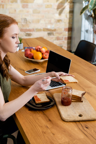 Widok boczny kobiety wylewanie dżemu na tosty w pobliżu urządzeń cyfrowych na stole  - Zdjęcie, obraz