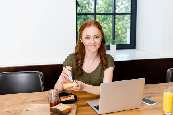 Kobieta uśmiecha się do kamery trzymając tosty z dżemem w pobliżu soku pomarańczowego i urządzeń cyfrowych na stole  - Zdjęcie, obraz