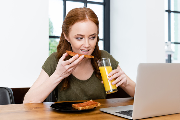Vrouw die toast eet en glas sinaasappelsap in de buurt van laptop op houten tafel houdt  - Foto, afbeelding