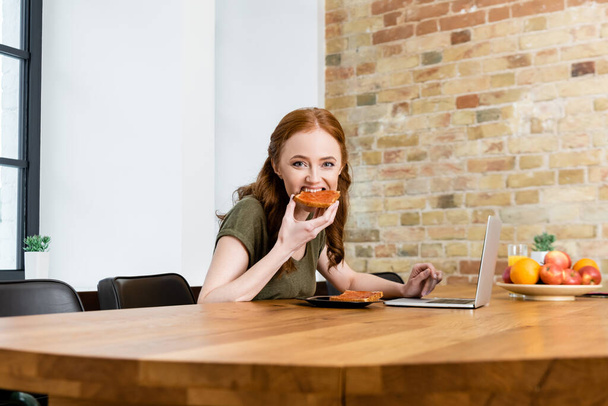 Selective focus of woman looking at camera while eating toast near laptop on table  - Photo, image