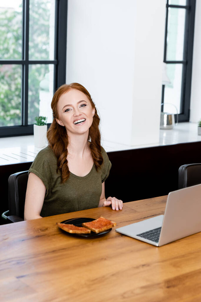 Attractive woman smiling at camera near toasts with jam and laptop on wooden table  - Photo, Image