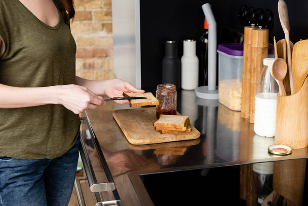 Cropped view of woman pouring jam on toast on kitchen worktop  - Photo, Image