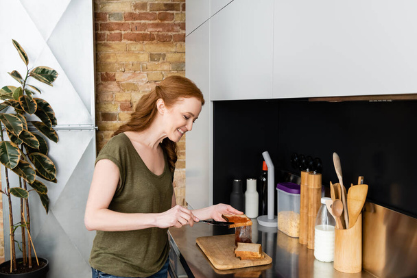 Smiling woman pouring jam on toast near kitchen worktop  - Foto, imagen