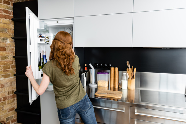 Πίσω όψη της γυναίκας που στέκεται κοντά σε ανοιχτό ψυγείο στην κουζίνα  - Φωτογραφία, εικόνα