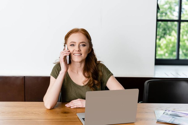 Smiling freelancer looking at camera while talking on smartphone near laptop on table  - Foto, Bild