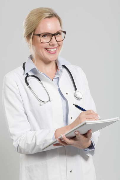 young friendly female doctor in doctor's overall with stethoscope takes notes for anamnesis - Photo, Image