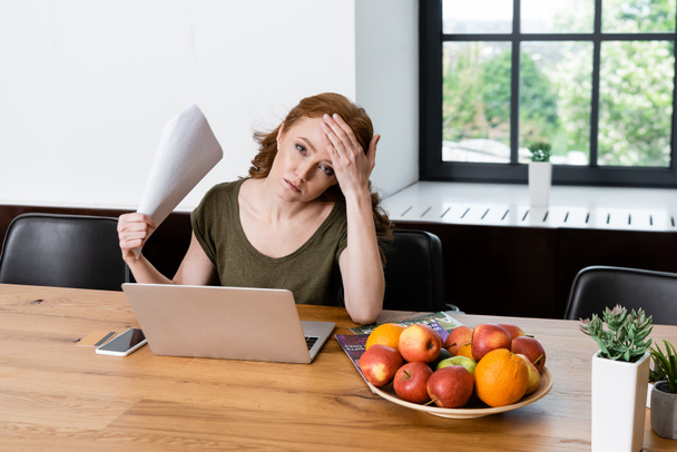 Woman holding paper while suffering from heat near gadgets and fruits on table  - Foto, imagen