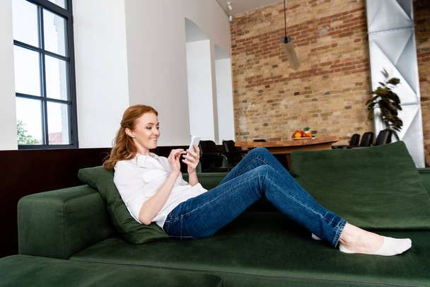 Vista lateral da mulher atraente sorrindo ao usar o smartphone no sofá
  - Foto, Imagem