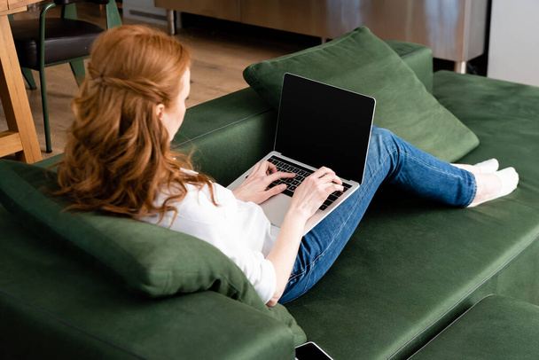 Szelektív fókusz vörös hajú szabadúszó a laptop közelében okostelefon kanapén  - Fotó, kép