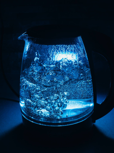 Boiling glass black teapot with blue backlight on a black background. Boiling water. Hot water is seething. Bubbles of air in the water. - Photo, Image