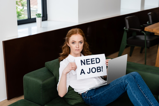 Attractive freelancer looking at camera while holding card with need a job lettering and laptop on couch  - 写真・画像