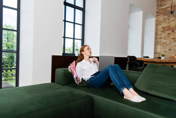 Приваблива жінка тримає журнал і ноутбук, страждаючи від спеки на дивані у вітальні
 - Фото, зображення