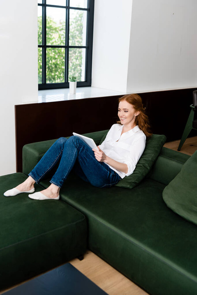 Femme souriante utilisant une tablette numérique tout en étant assis sur le canapé dans le salon  - Photo, image