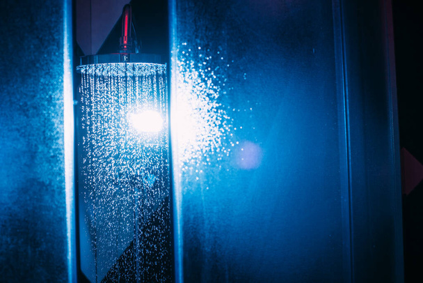 Gotas de agua caen de una regadera en la ducha con luz azul. Gotas de agua en primer plano. Flujo de agua. Una gran regadera redonda en la ducha
. - Foto, Imagen