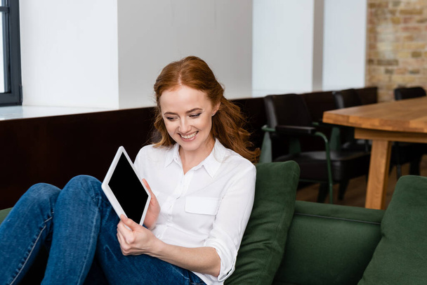 Lächelnde Frau zeigt digitales Tablet mit leerem Bildschirm, während sie zu Hause auf der Couch sitzt  - Foto, Bild