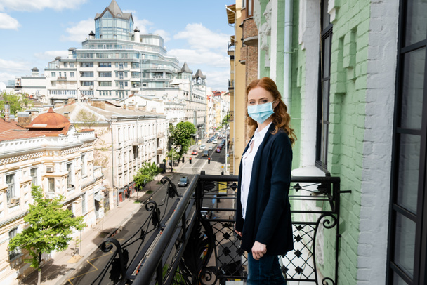 Zijaanzicht van vrouw in medisch masker kijkend naar camera op terras  - Foto, afbeelding