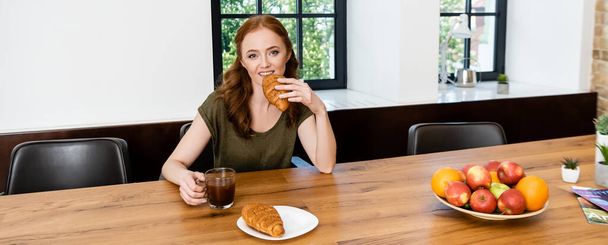 Panoramic orientation of woman eating croissant and holding cup of coffee during breakfast  - Photo, Image