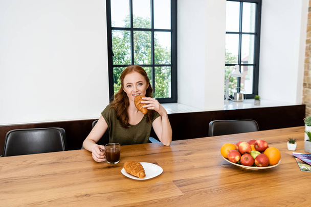 Red haired woman eating croissant holding cup of coffee at home  - Fotó, kép
