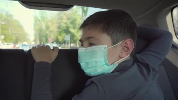 Menino triste em máscara protetora no banco de trás do carro em movimento no trânsito da cidade durante a epidemia de coronavírus e auto-isolamento
 - Filmagem, Vídeo