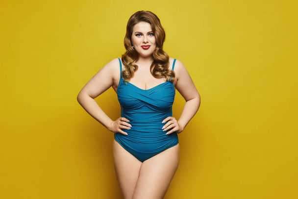 Plus-size model girl with stylish hairstyle and red lips in fashionable swimsuit at the yellow background, isolated. Fat woman in swimsuit on yellow background. - Фото, изображение