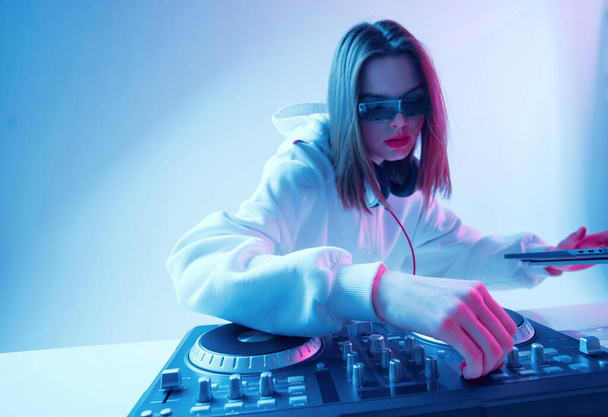 Cool young girl DJ mixes music on a mixing console and laptop, in stylish clothes, glasses on a neon background. - Фото, зображення