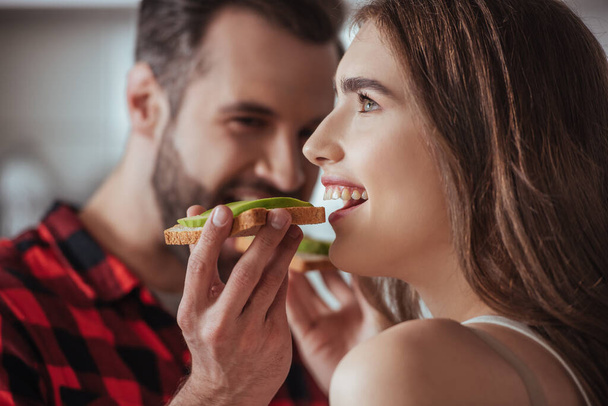 selective focus of smiling man feeding happy girlfriend with fresh avocado toast  - Fotó, kép